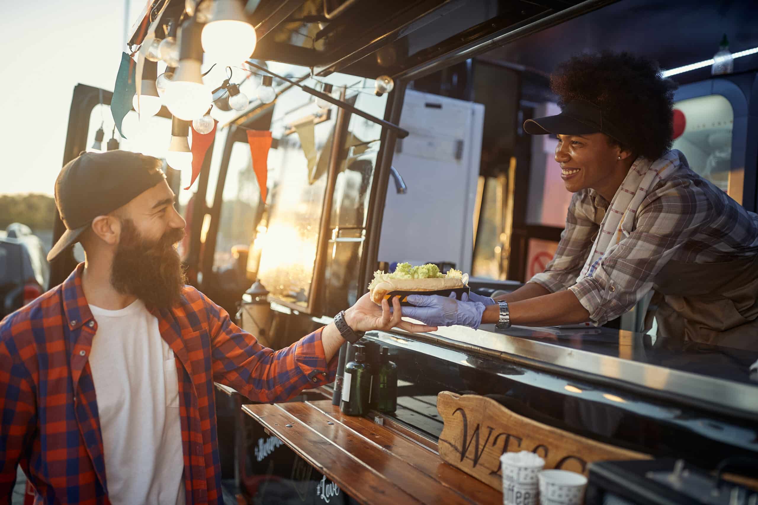 afro-american female employee gives with care and smile a sandwich to young hipster male customer